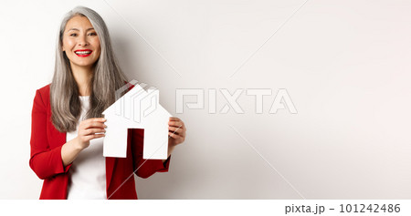 Asian female real estate agent showing paper house cutout, broker smiling friendly and selling property, standing over white background 101242486