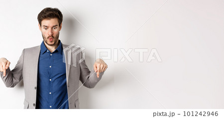 Wow look at this. Impressed and excited businessman in suit pointing fingers down, looking at bottom advertisement with amazed face, standing on white background 101242946