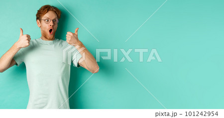 Excited young man with red hair and beard, staring at promo with awe, showing thumbs-up in approval, praising offer, standing over turquoise background 101242954