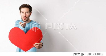 Romantic boyfriend sending air kiss at camera and holding big red Valentines day heart, standing over white background 101243012