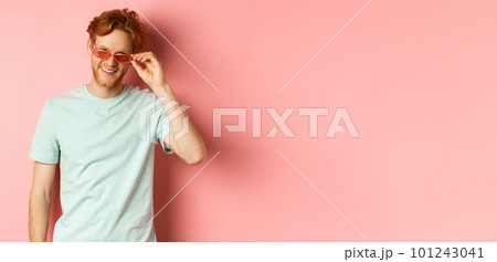 Tourism and vacation concept. Relaxed young man with red hair, standing in sunglasses and t-shirt and smiling pleased, standing over pink background 101243041