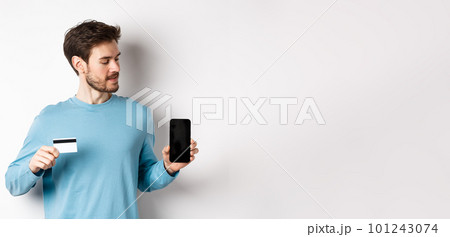 Mobile banking. Handsome caucasian man showing plastic credit card and looking at blank mobile screen, standing over white background 101243074