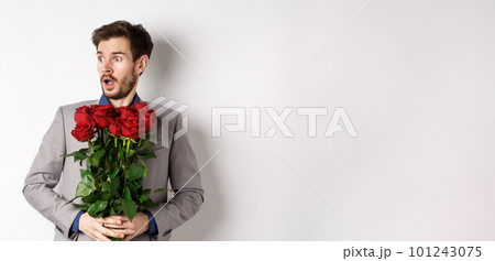 Handsome young man in suit holding red roses, looking left with surprised and startled expression, standing on Valentines day over white background 101243075