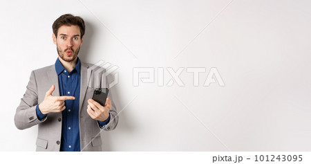 Excited guy in suit say wow, found something online on smartphone, pointing at mobile phone impressed, white background 101243095