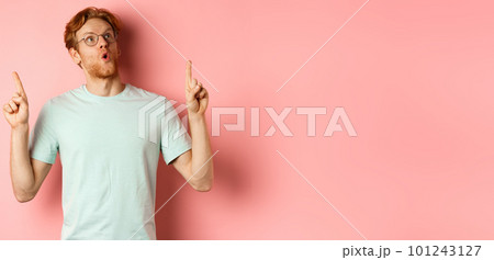Wow check this out. Surprised redhead guy in glasses looking and pointing up, gasping amazed, standing over pink background 101243127