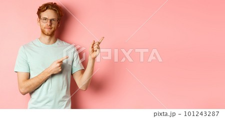 Confident young man with ginger hair and beard, smiling pleased and pointing fingers at upper right corner, invite or demonstrate something, standing over pink background 101243287