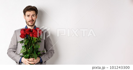 Worried boyfriend in suit, holding flowers roses and looking doubtful at camera, standing with bouquet on valentines day against white background 101243300