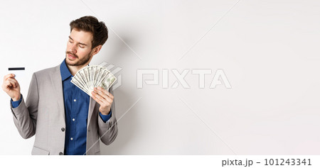 Successful man holding dollar bills and looking at plastic credit card, pleased with bank, standing on white background in business suit 101243341