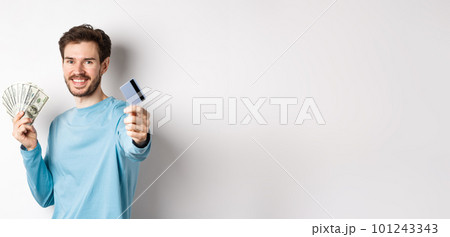 Smiling caucasian man holding money and giving you plastic credit card, standing on white background 101243343