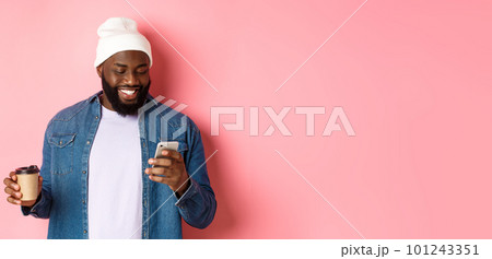 Image of stylish Black man hipster drinking takeaway coffee, reading message on phone and smiling, standing over pink background 101243351