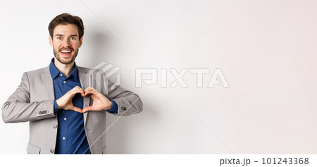 Passionate and romantic man in suit falling in love, showing heart sign and smiling at lover, standing on white background 101243368