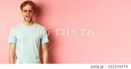 Image of surprised redhead man on vacation, wearing sunglasses with summer t-shirt, open mouth and saying wow amazed, standing over pink background 101243374