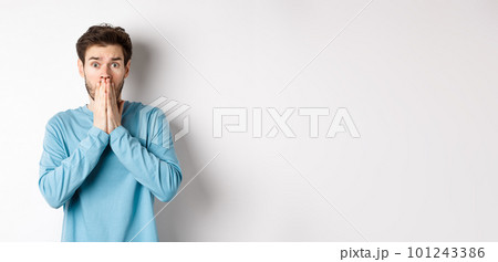 Shocked and worried young man gasping and covering mouth, look with panic at camera, standing anxious against white background Shocked and worried young man gasping and covering mouth, look with panic at camera, standing anxious against white background 101243386