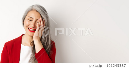 Beauty and makeup concept. Close up of beautiful asian mature woman, laughing and touching face, smiling happy, standing over white background 101243387