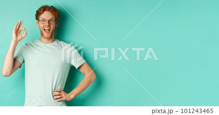 Amused young man with red hair, wearing glasses and t-shirt, showing okay sign and smiling excited, checking out something and approving it, standing over turquoise background Amused young man with red hair, wearing glasses and t-shirt, showing okay sign and smiling excited, checking out something and approving it, standing over turquoise background 101243465