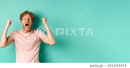 Handsome redhead man watching sports game and cheering, shouting and making fist pumps, looking at screen and watching competition, rooting for team, standing over turquoise background 101243473