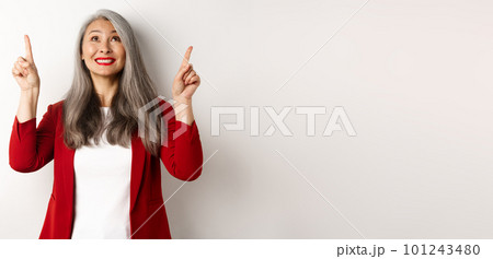 Happy asian lady in red blazer and makeup, looking and pointing fingers up, showing special offer, standing over white background Happy asian lady in red blazer and makeup, looking and pointing fingers up, showing special offer, standing over white background 101243480