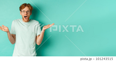 Surprised handsome man with red hair, wearing glasses, spread hands sideways and gasping in awe, looking wondered at camera, standing over turquoise background 101243515