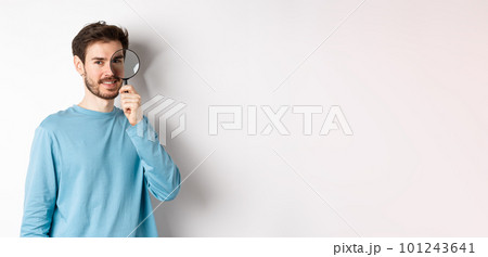 Smiling young man detective looking through magnifying glass, searching for promo offer, standing on white background Smiling young man detective looking through magnifying glass, searching for promo offer, standing on white background 101243641