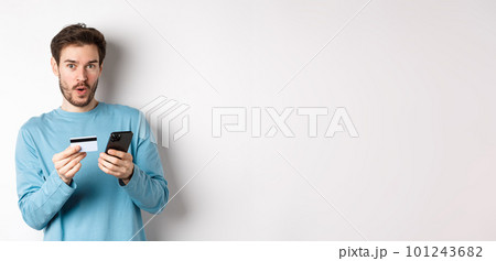 E-commerce and shopping concept. Excited man looking amused while making order online, holding plastic credit card and smartphone, standing on white background E-commerce and shopping concept. Excited man looking amused while making order online, holding plastic credit card and smartphone, standing on white background 101243682