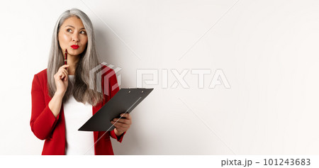 Stylish asian office lady in red blazer writing report on clipboard, looking thoughtful while working, standing over white background 101243683