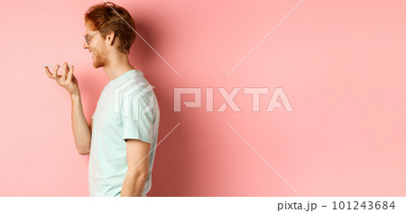 Profile portrait young man with red hair, smiling pleased while record voice message on smartphone, talking to virtual assistant, standing over pink background 101243684