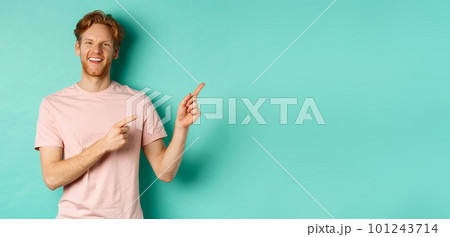 Handsome male model with red messy hair showing advertisement on copy space, pointing at upper right corner and smiling happy, standing over turquoise background 101243714