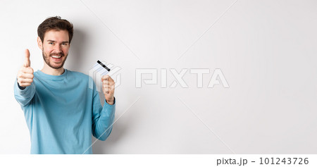 Cheerful young man showing plastic credit card and thumb up, like and approve good bank service, standing over white background Cheerful young man showing plastic credit card and thumb up, like and approve good bank service, standing over white background 101243726