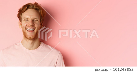 Close up of cheerful young man with messy red hair and beard, laughing and smiling, showing white teeth, looking happy at camera, standing over pink background 101243852
