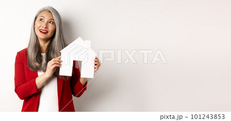 Dreamy senior woman thinking of buying property, showing paper house cutout and looking at upper left corner, standing over white background Dreamy senior woman thinking of buying property, showing paper house cutout and looking at upper left corner, standing over white background 101243853