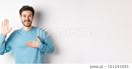 Smiling hosent man raising arm and hold hand on heart, making promise tell truth, swear or give oath, standing over white background 101243893