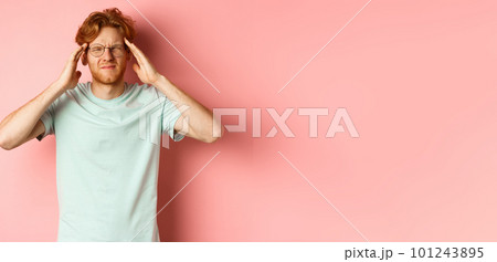Portrait of redhead man in crooked glasses touching head and feeling dizzy or nauseous, having hangover or headache, standing over pink background 101243895