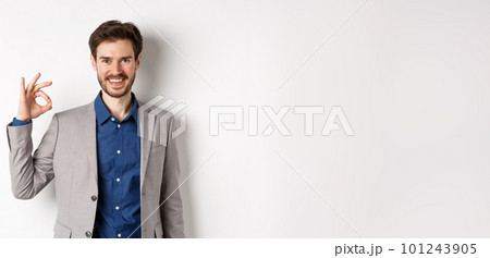 Smiling successful businessman in suit showing okay sign, looking satisfied, standing against white background Smiling successful businessman in suit showing okay sign, looking satisfied, standing against white background 101243905
