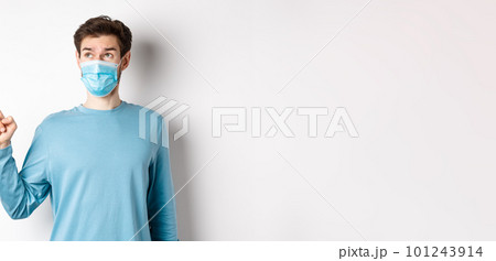 Coronavirus, health and quarantine concept. Curious guy in medical mask, pointing and looking at upper left corner banner, standing on white studio background Coronavirus, health and quarantine concept. Curious guy in medical mask, pointing and looking at upper left corner banner, standing on white studio background 101243914