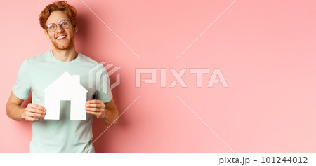 Real estate. Cheerful young man with red hair, wearing glasses and t-shirt, showing paper house cutout and smiling, buying apartment, pink background 101244012