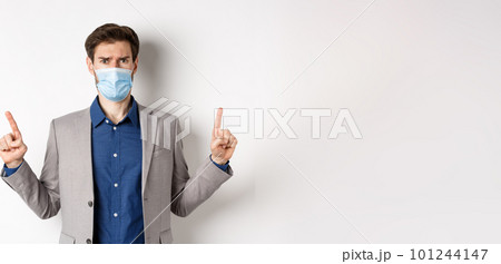 Covid-19, pandemic and business concept. Disappointed frowning guy in suit and medical mask pointing fingers up, complaining on unfair situation, white background 101244147
