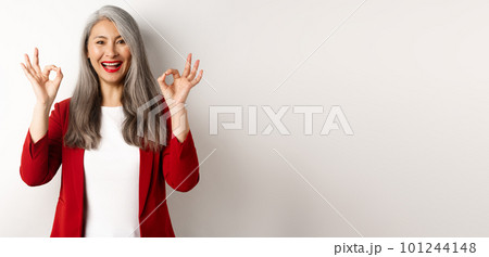 Beautiful senior asian woman in red blazer, smiling and showing okay signs, approve and say yes, standing over white background 101244148