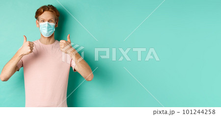 Covid-19, pandemic and lifestyle concept. Cheerful redhead guy in medical mask showing thumbs up in approval, like and praise product, standing over turquoise background Covid-19, pandemic and lifestyle concept. Cheerful redhead guy in medical mask showing thumbs up in approval, like and praise product, standing over turquoise background 101244258