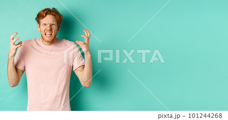 Annoyed and pissed-off redhead guy looking angry, shouting and grimacing, shaking hands aggressive, standing furious against mint background 101244268