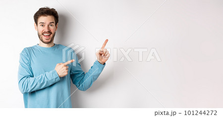 Cheerful smiling man pointing fingers right and showing banner or logo on white space for advertisements, recommending product, standing over studio background 101244272