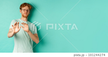 Reluctant redhead man in glasses raising hands in refusal gesture, step backwards, avoiding something with disgusted and unamused face, standing over turquoise background 101244274