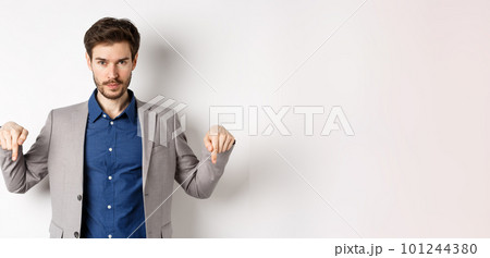 Look here. Handsome bearded man in suit pointing fingers down, looking confident at camera, showing banner, standing on white background Look here. Handsome bearded man in suit pointing fingers down, looking confident at camera, showing banner, standing on white background 101244380