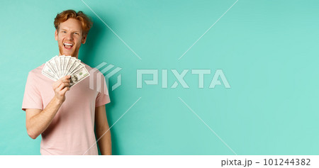 Happy redhead man in t-shirt showing money in dollars and smiling, making smug faces after winning cash, standing over turquoise background 101244382
