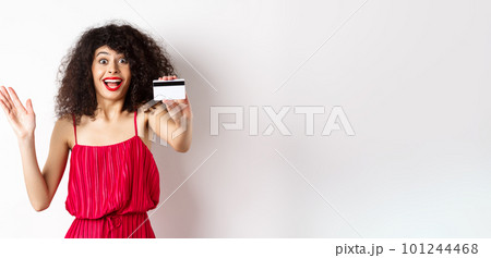 Shopping. Amazed curly-haired woman in red dress, showing plastic credit card and scream from excitement, standing on white background 101244468