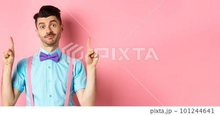 Cheerful funny man in bow-tie pointing fingers up, showing advertisement and smiling indecisive, standing over pink background Cheerful funny man in bow-tie pointing fingers up, showing advertisement and smiling indecisive, standing over pink background 101244641
