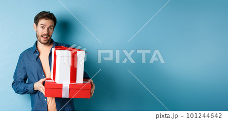 Handsome bearded guy holding Valentines day gifts for lover, standing with presents in boxes and looking at camera, blue background Handsome bearded guy holding Valentines day gifts for lover, standing with presents in boxes and looking at camera, blue background 101244642
