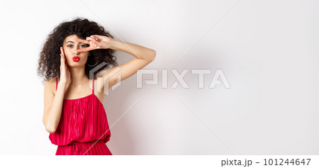 Beauty and fashion. Stylish curly-haired woman in red dress showing v-sign on eye and pucker lips, kissing you, standing on white background Beauty and fashion. Stylish curly-haired woman in red dress showing v-sign on eye and pucker lips, kissing you, standing on white background 101244647
