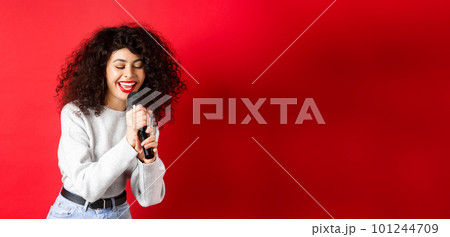 Hobbies and leisure concept. Beautiful woman singing in microphone and smiling, having fun at karaoke, standing on red background 101244709