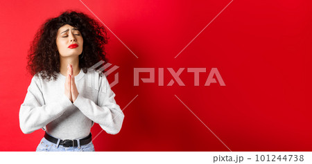 Desperate young woman pleading God, making wish and supplicating, holding hands in pray with eyes close and begging face, standing on red background 101244738