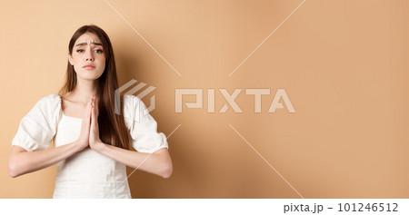 Please help. Desperate young woman begging you, holding hands in pleading gesture and looking sad at camera, asking for favour, beige background 101246512